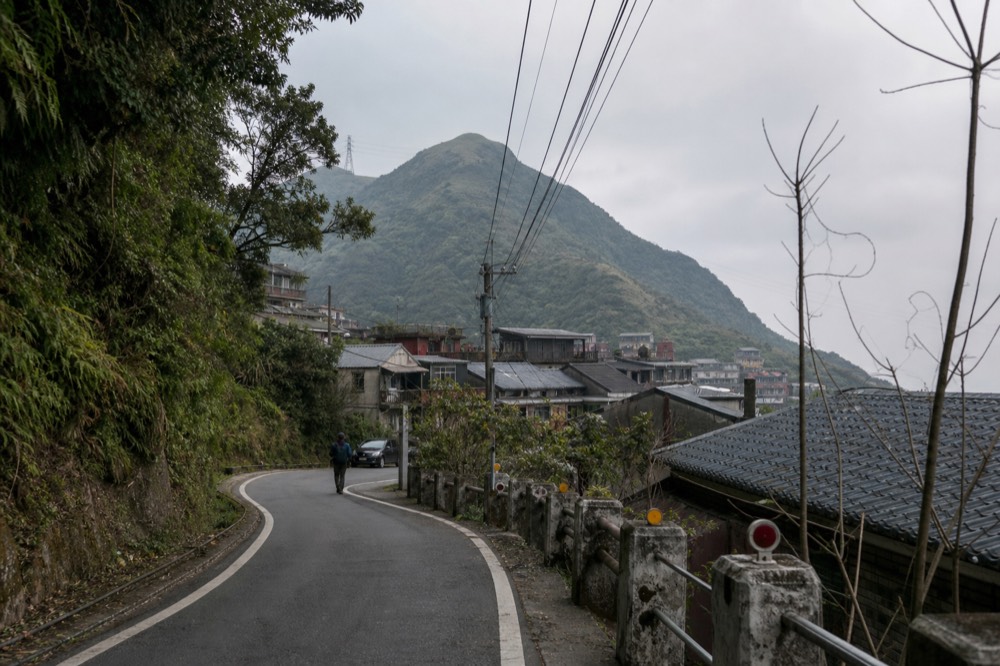 A winding mountain road climbing a forested hillside toward hillside rooftops under an overcast sky
