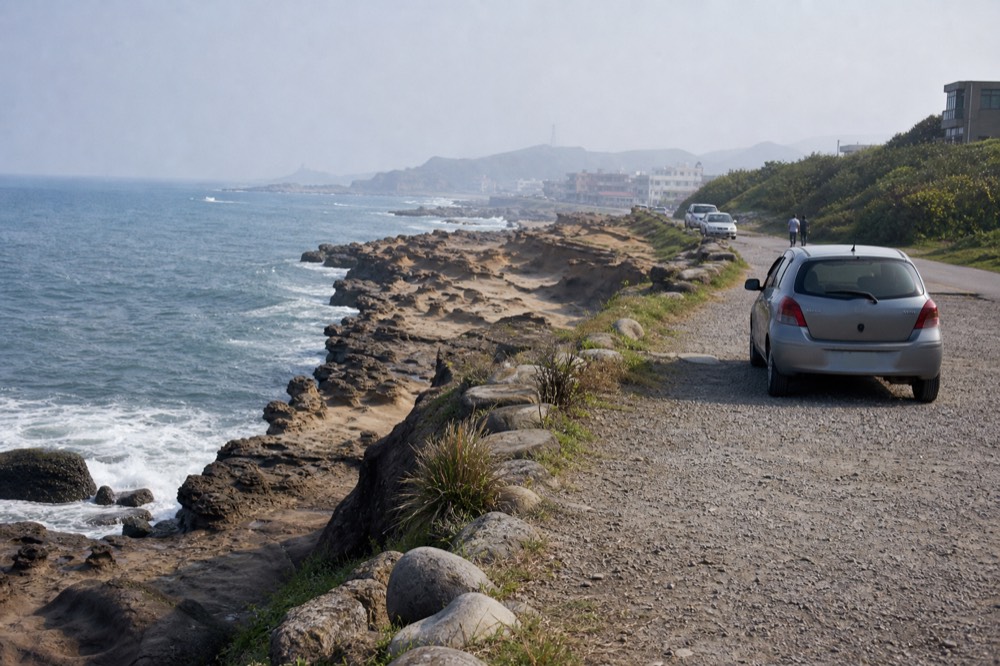 A coastal road curving along a rocky shoreline with waves breaking against dark stone