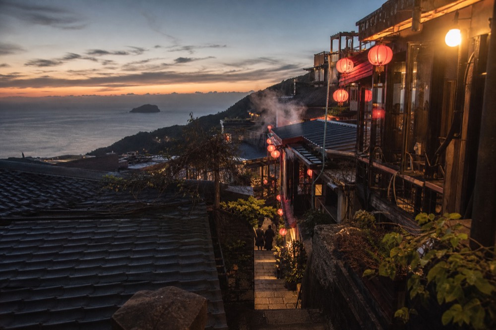 A hillside town at dusk with warm lantern light glowing along stacked rooftops above the sea