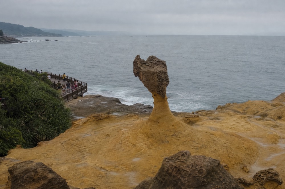 A tall slender mushroom rock formation rising from an eroded coastal platform with the sea behind