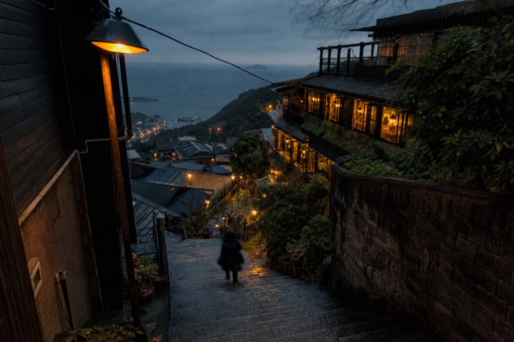 A hillside town of lit lanterns and old buildings above a dark sea at dusk