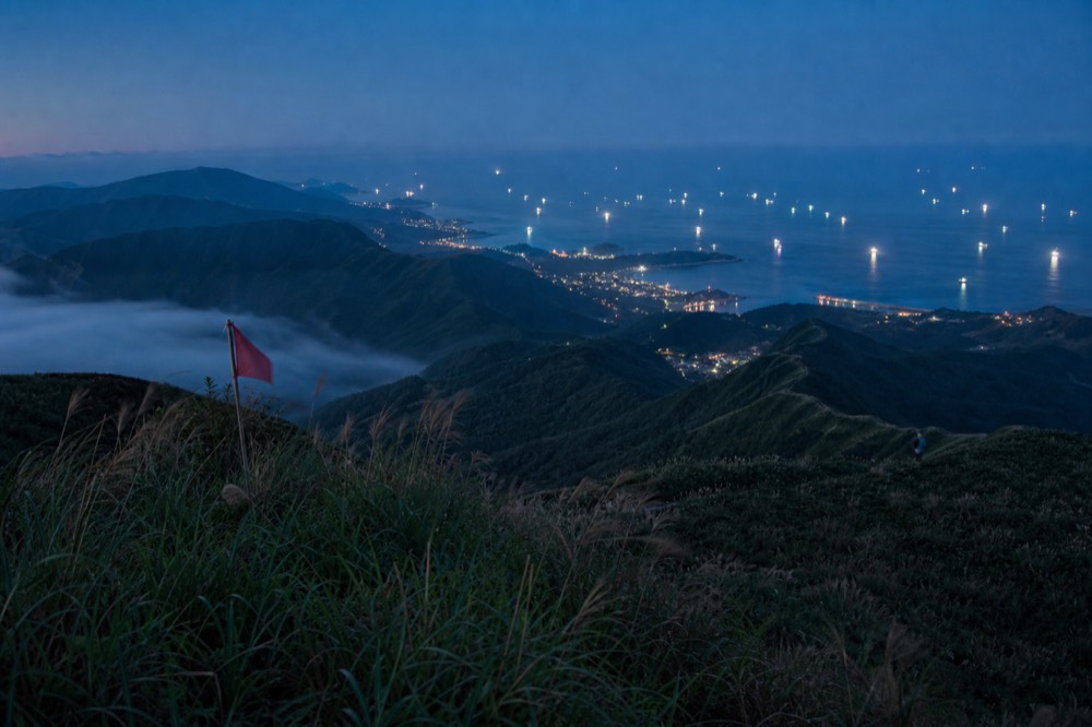 Distant coastline lights below a dark hillside town under an evening sky