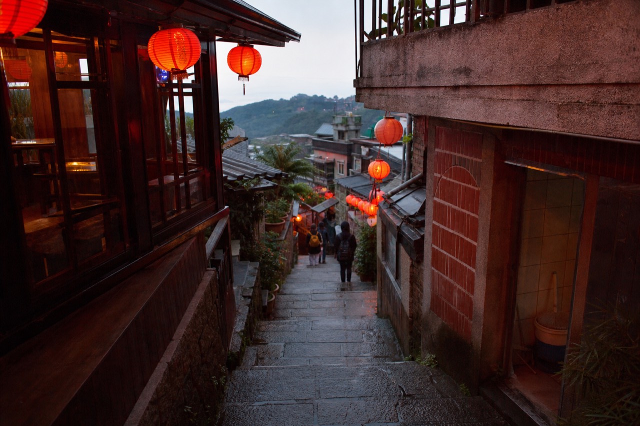 Red lanterns glowing along a narrow stepped stone lane in a hillside town at dusk