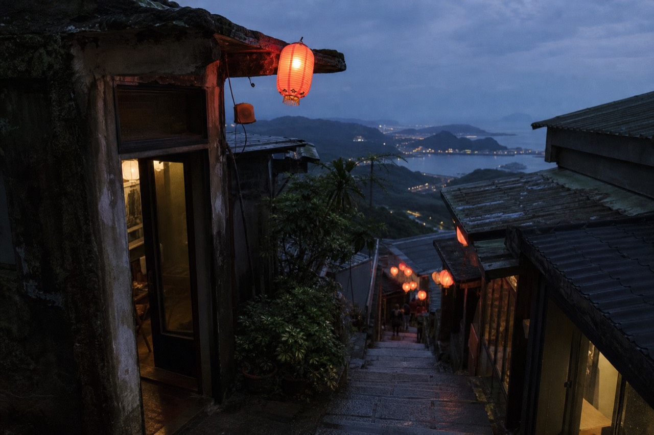Lantern-lit stone steps of a hillside old street descending toward a distant coastline at dusk