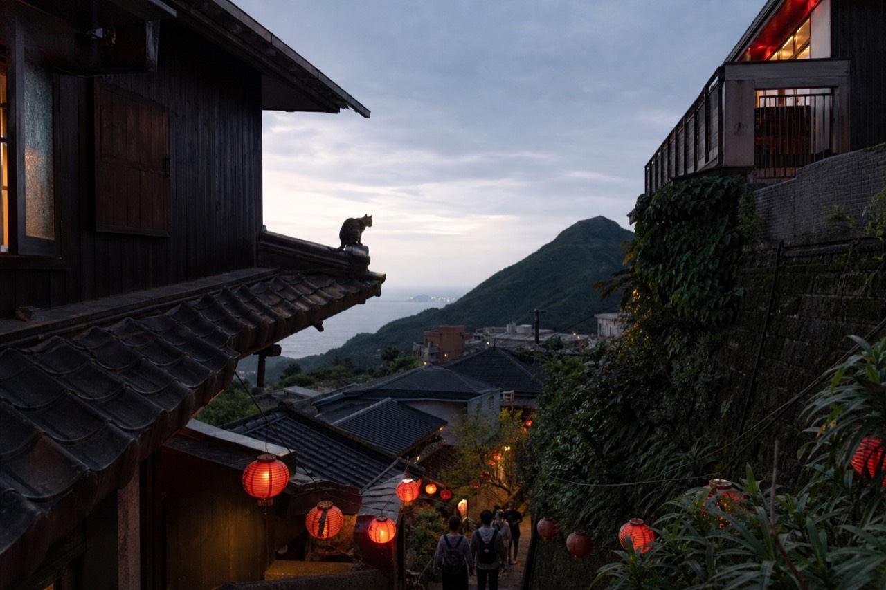 A hillside old street with red lanterns and tiled rooftops descending toward a distant coastline