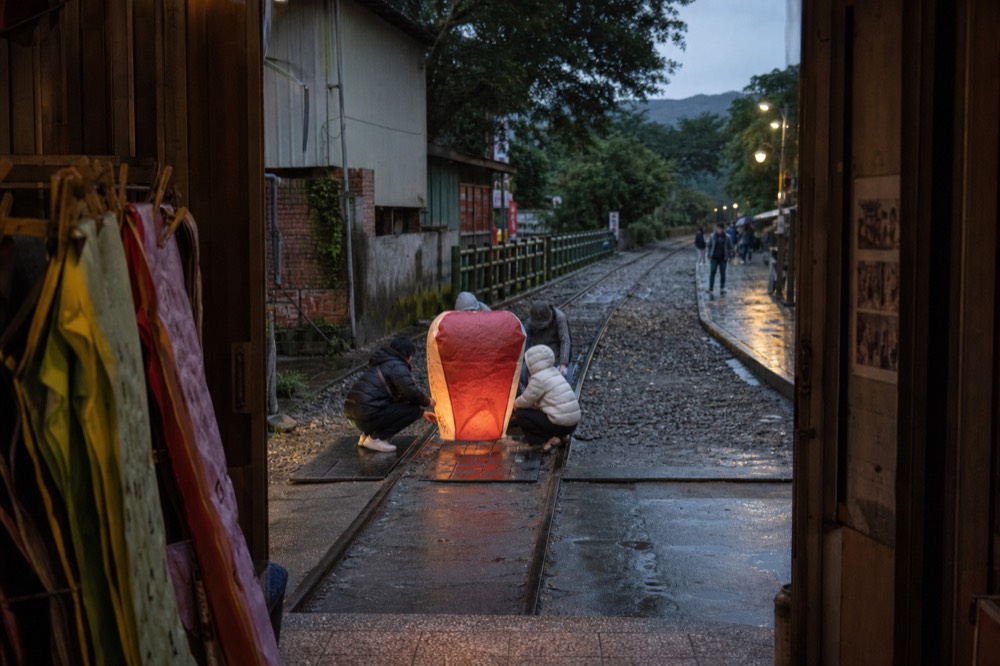 A crowd of visitors releasing a glowing paper lantern beside a railway track in a narrow valley town