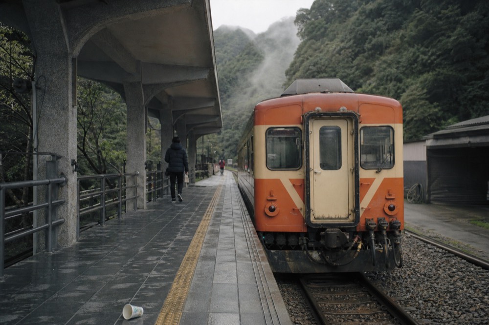 A single-car diesel train waiting at a small rural station platform in a wooded valley