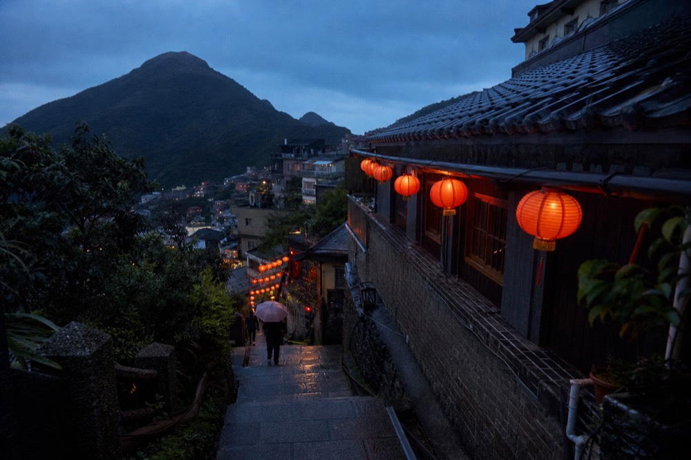Red lanterns glowing along the eaves of a steep old street at dusk with hills behind