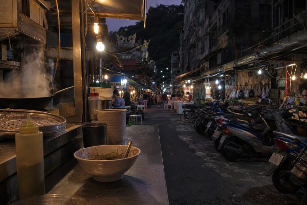 Food stalls with hanging yellow lights along a crowded narrow lane in a Taiwanese port city at night