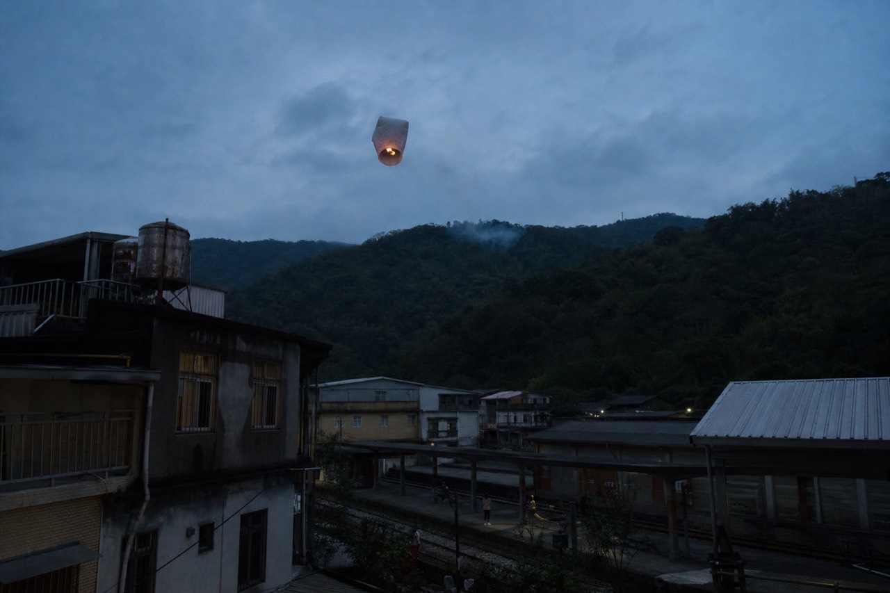 A sky lantern drifting above the rooftops and hills of a mountain town at dusk