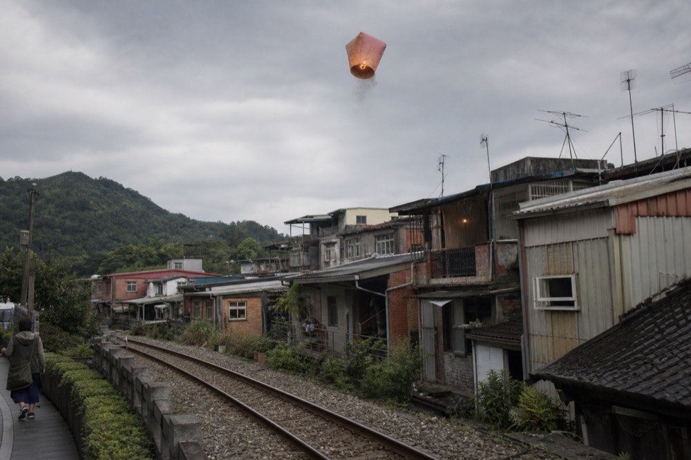 A sky lantern rising above rooftops beside railway tracks in a small mountain town
