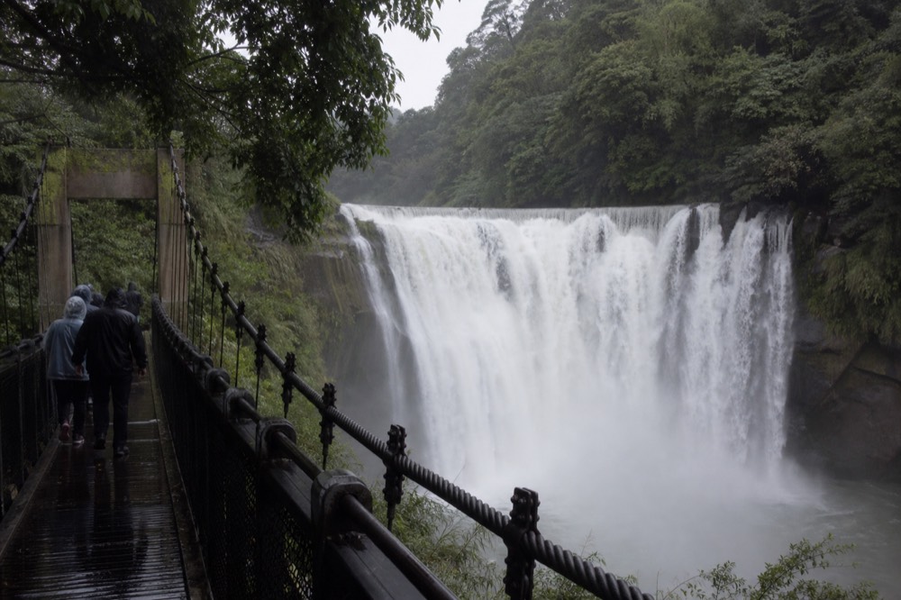 A wide curtain of water falling across a rock face with forested hills on both sides