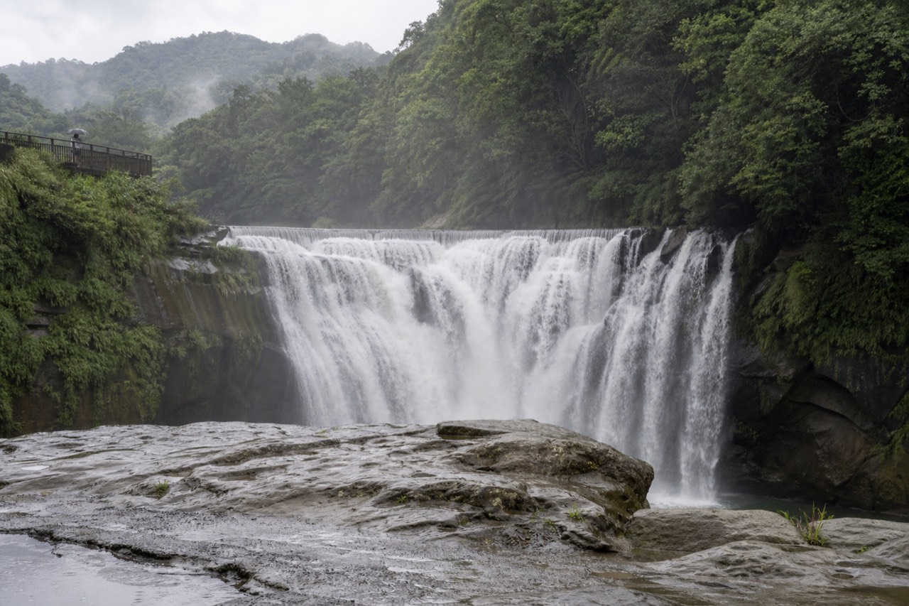 A waterfall pouring over a wide rock face above a small riverside town in the mountains of northern Taiwan