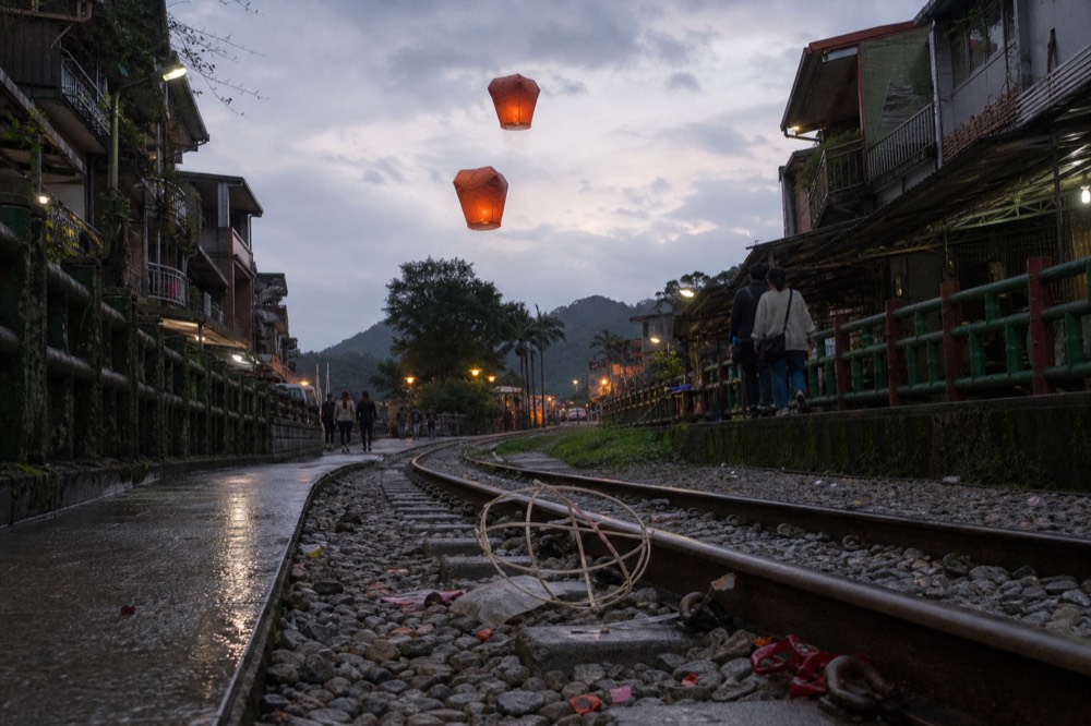 Red and orange sky lanterns rising above a railway line running between old street houses in a mountain valley town.