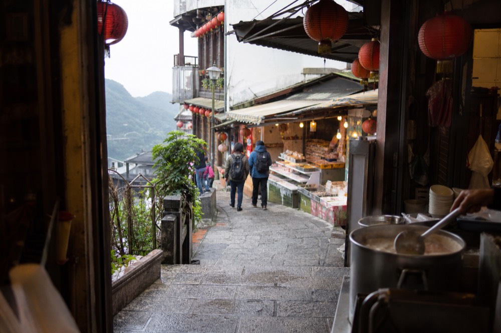 A narrow old street lined with red lanterns and snack stalls curving along a hillside above the sea in a former mining town.