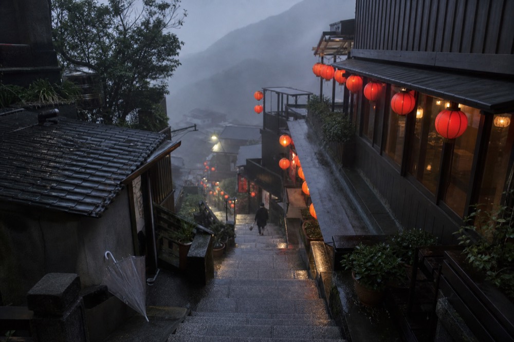 A steep lane of stone steps between old houses, with red lanterns glowing softly in drifting mist on a hillside above the sea.