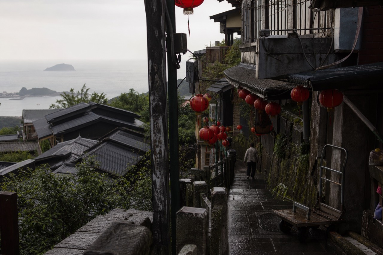 A hillside mining town with red lanterns and tiled rooftops descending toward a grey sea coastline under an overcast sky.
