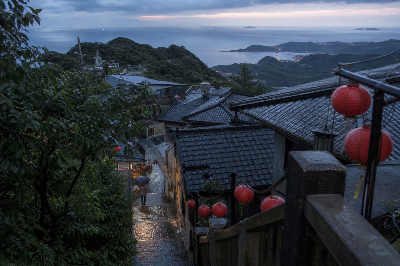 Lantern-lit rooftops of a hillside town above the sea under clearing skies