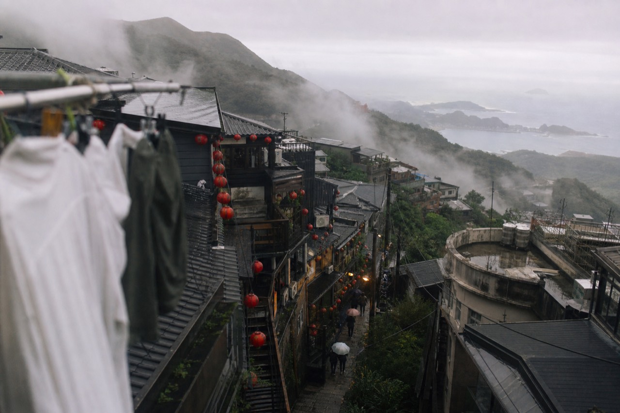 Steep hillside lanes with lanterns and old rooftops descending toward the sea in a foggy mountain town