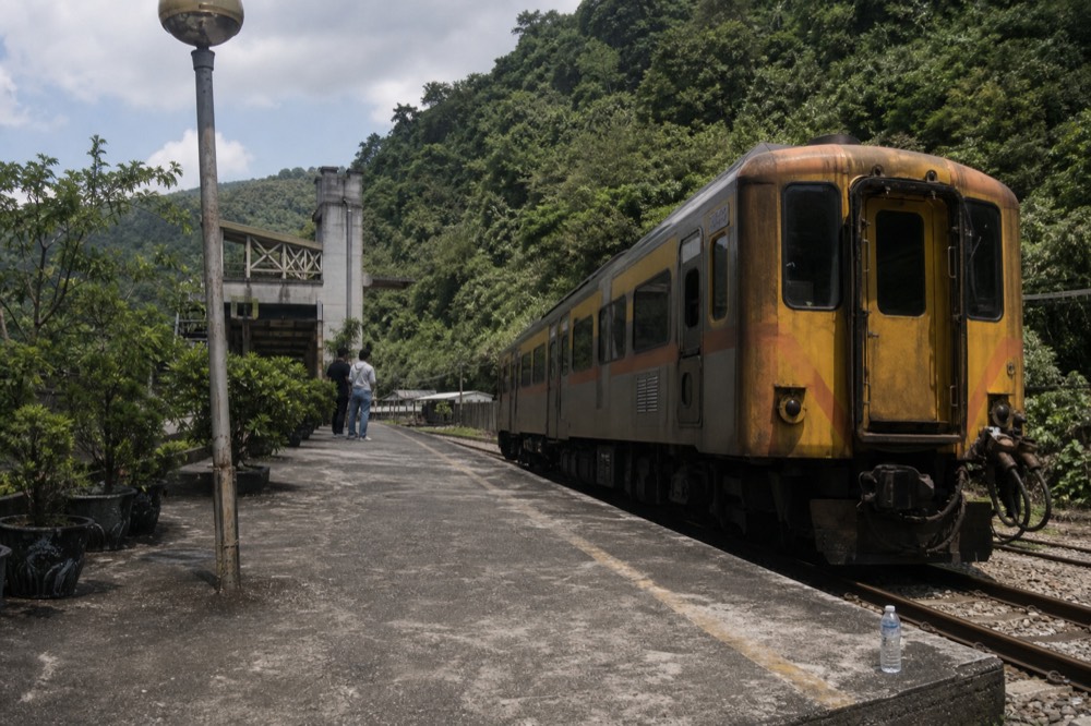 A rural branch line train waiting at a small station platform beside a river and forested hillside