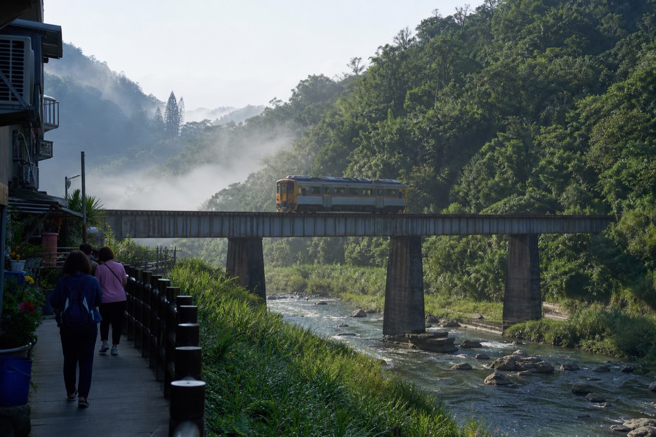 A single-carriage train crossing a railway bridge over a river between forested hills in rural Taiwan