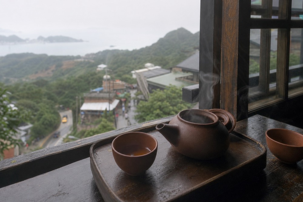 A tea set on a wooden table beside a window overlooking a misty hillside, with a pot of oolong tea and two small cups.
