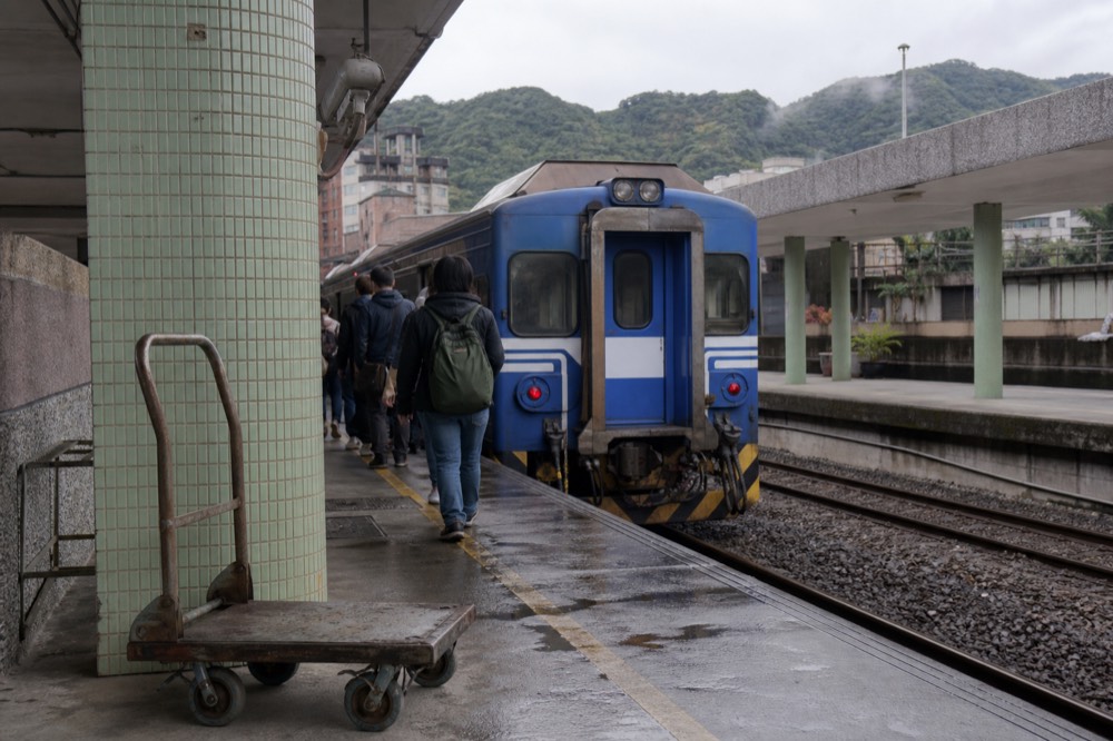 A train platform at Ruifang Station with a blue local train waiting, mountains visible in the background under overcast skies.