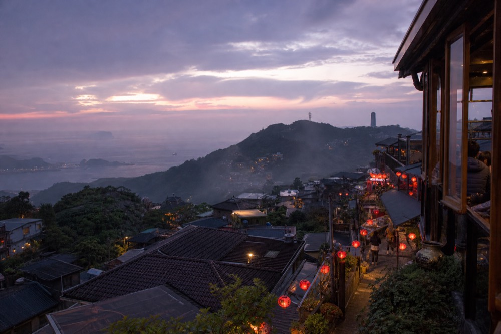 A view from a teahouse balcony at dusk, showing the hillside dotted with red lanterns and the sea in the distance under a fading sky.