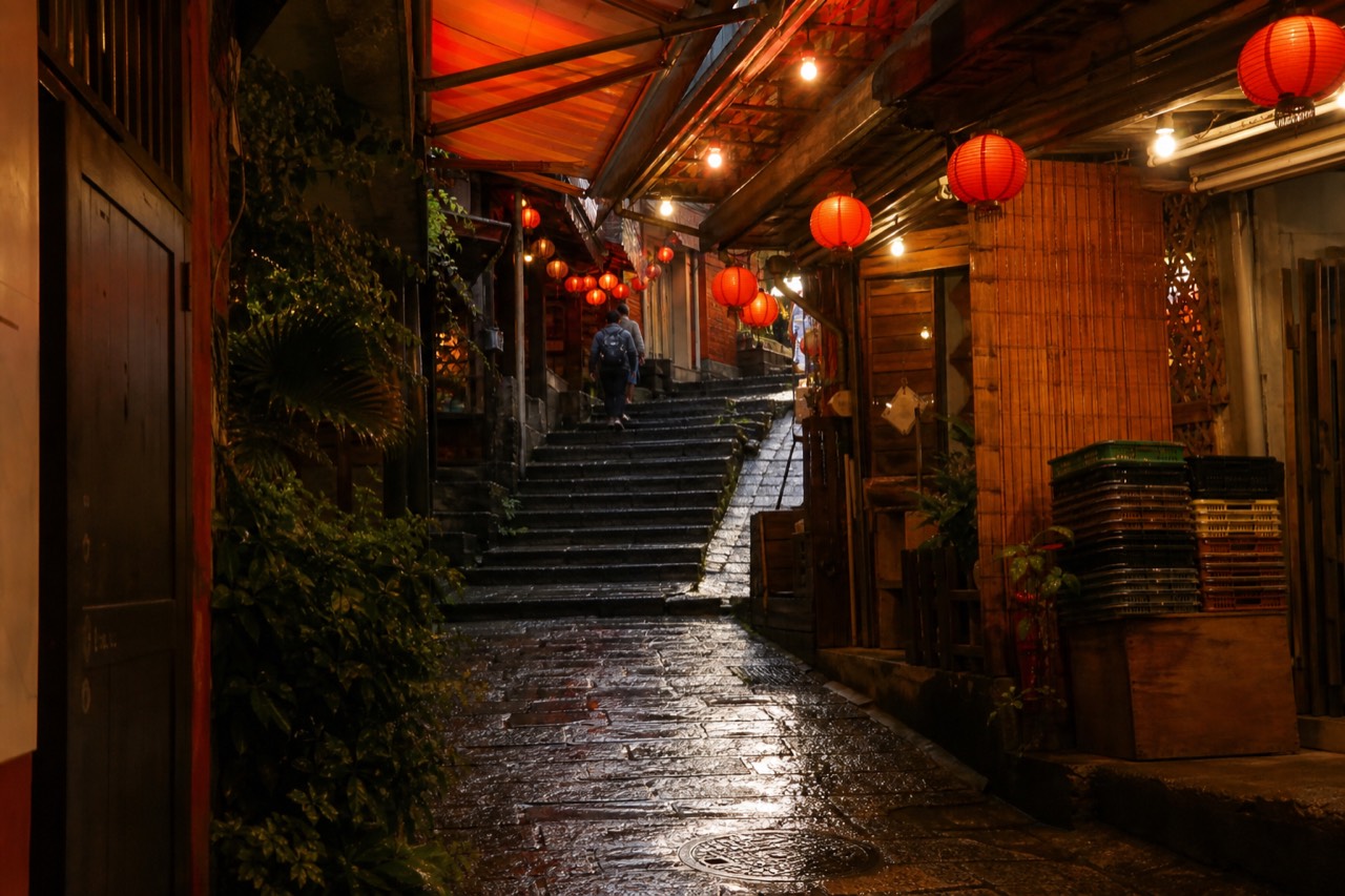 A narrow, lantern-lit alleyway in Jiufen Old Street during the evening, with red lanterns glowing and stone steps leading up between traditional shopfronts.