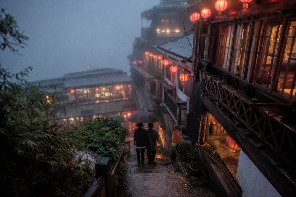 Steep stone steps between old teahouse buildings under red lanterns on a foggy hillside evening