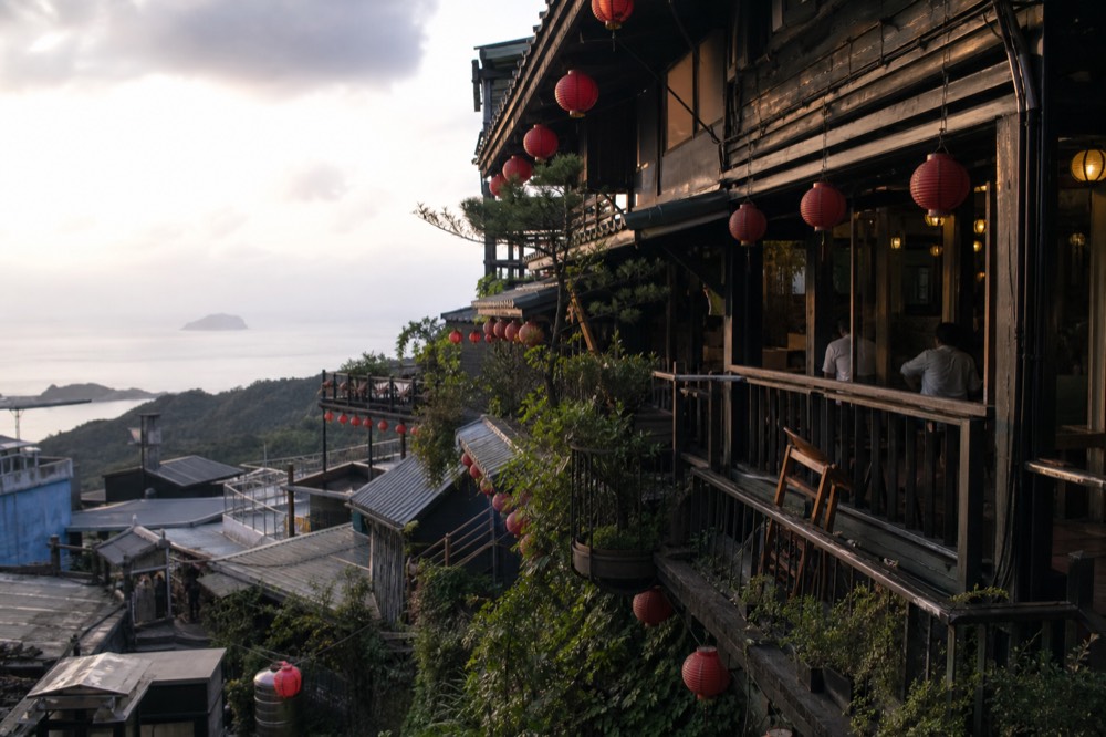 Wooden teahouse balconies stacked on a steep slope above the sea, with lanterns and tiled roofs
