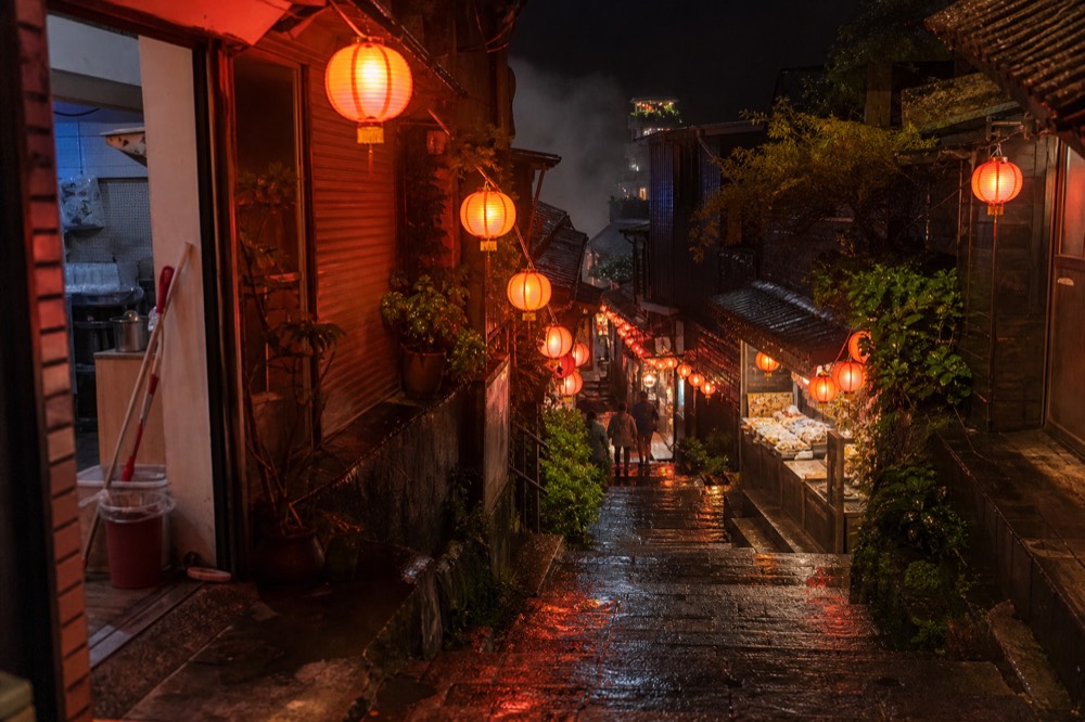 Red lanterns glowing over a wet stone lane at night, with lit shopfronts and mist above the rooftops