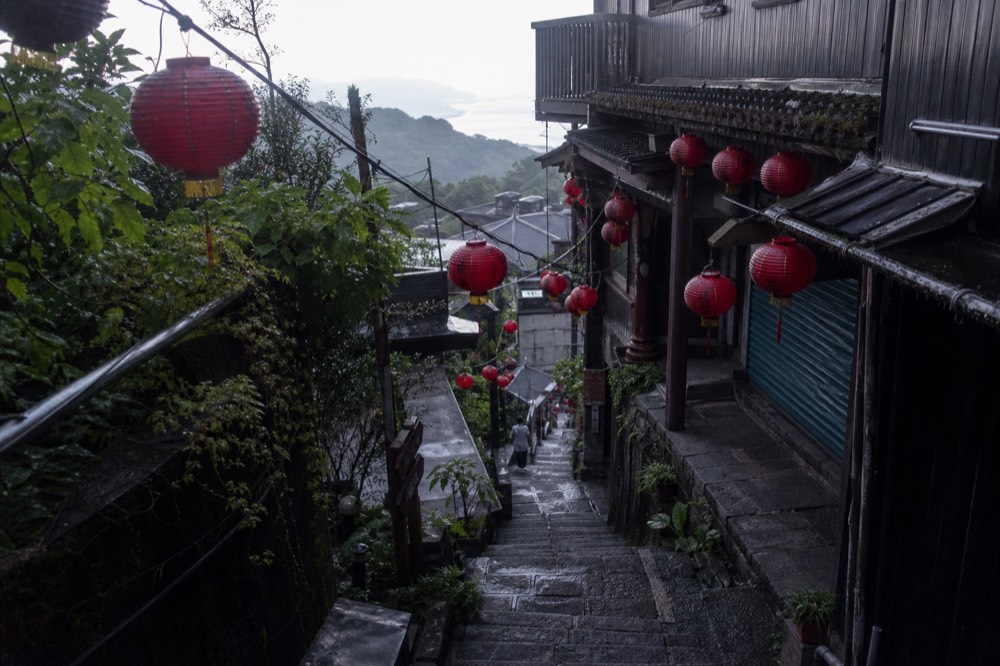 A narrow hillside lane with stone steps, teahouse signs and red lanterns, quiet in early morning light