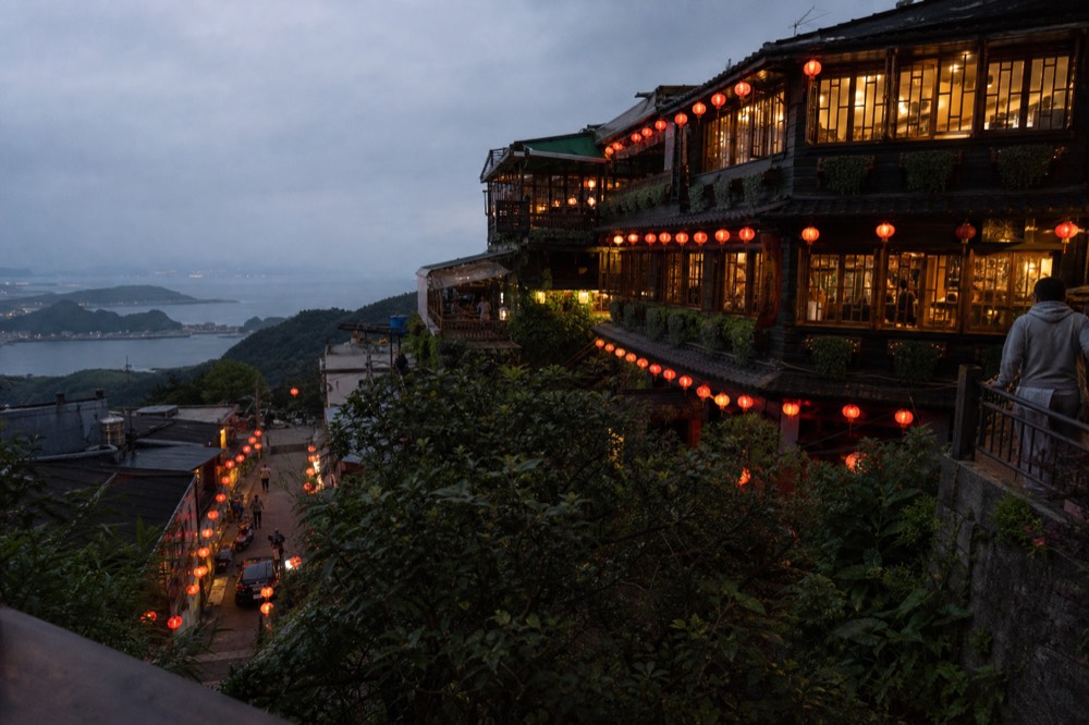 Red paper lanterns glowing along a narrow lane at dusk with hills and sea in the distance
