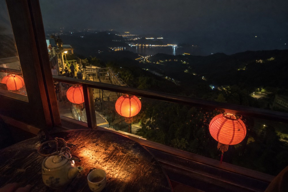 A wooden teahouse balcony with lit lanterns overlooking dark hills and a distant coastline at night