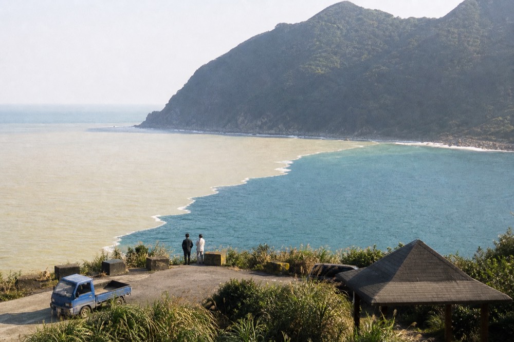 A coastal bay where yellow-tinted water meets blue sea below a forested headland