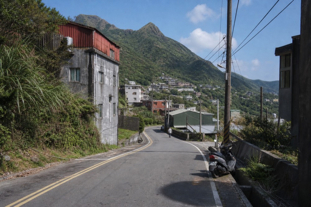 A narrow mountain road winding between a hillside village and a coastal valley