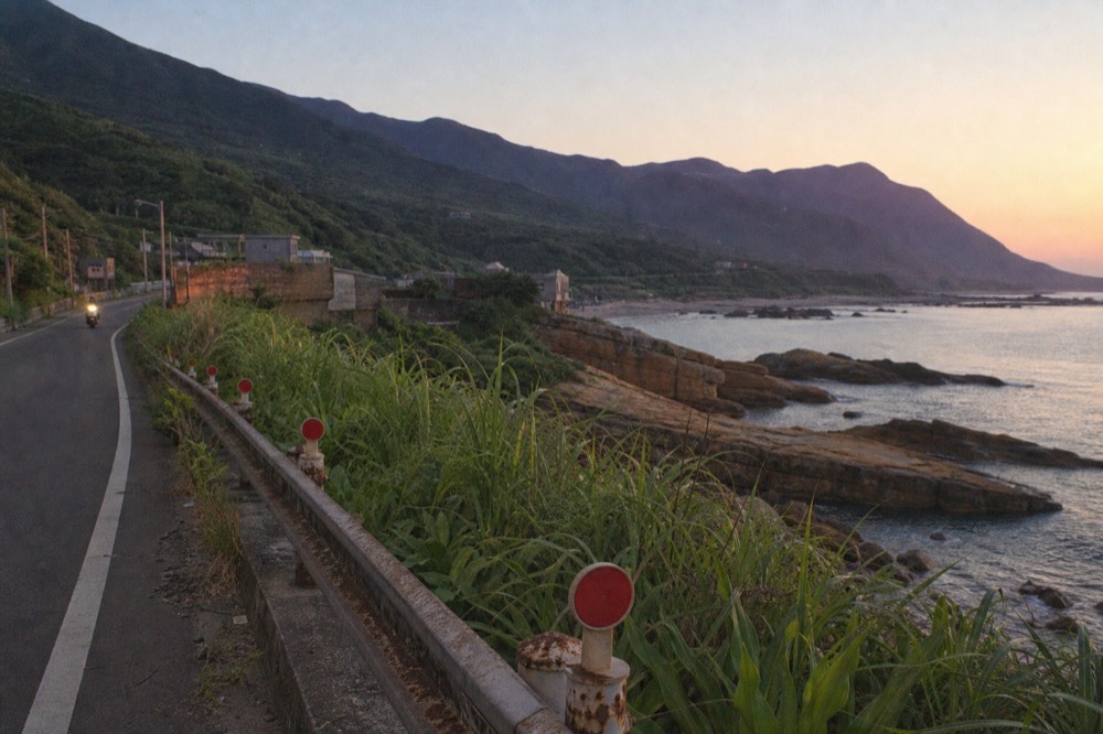 A coastal road at dusk with orange-stained rocks and a calm sea below hills