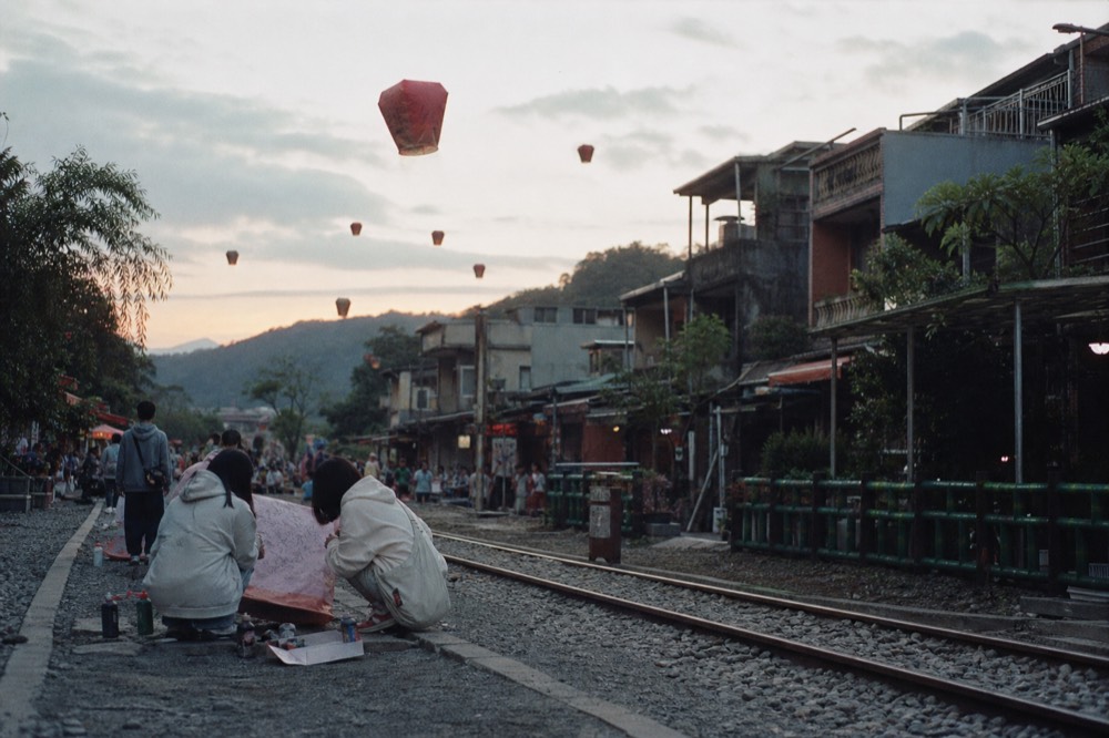 Sky lanterns rising above Shifen railway tracks, with a crowd of visitors painting lanterns on the pavement