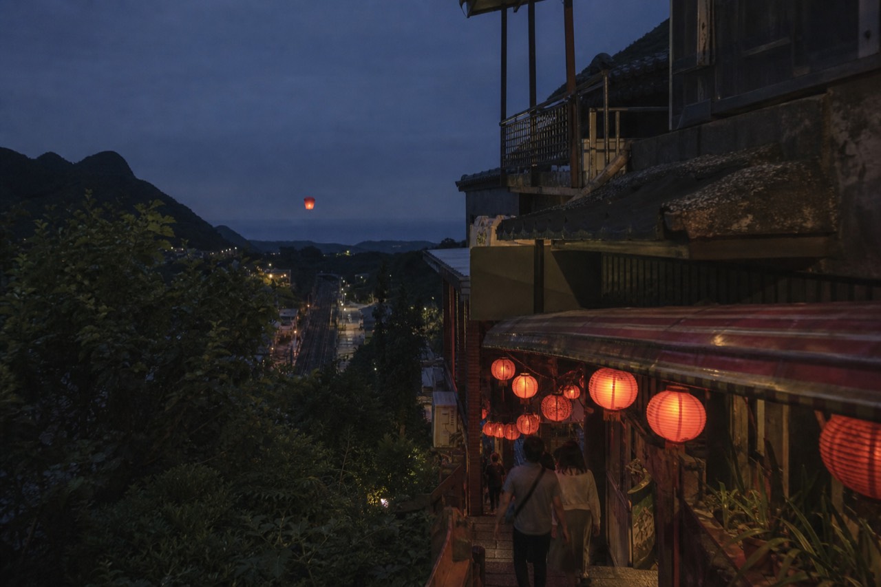 View of red lanterns hanging over Jiufen Old Street at dusk, with a sky lantern rising over Shifen's railway tracks in the distance