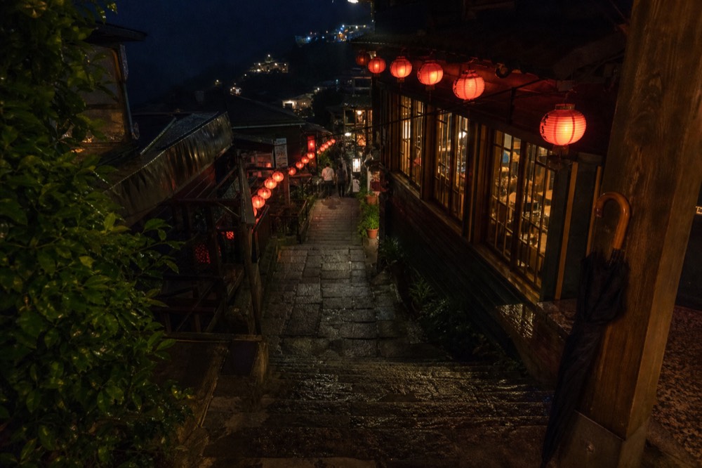 A narrow stone street lit by rows of red lanterns at dusk with teahouse windows glowing