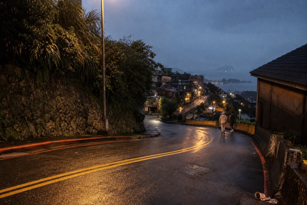 A hillside road above a coastal town in late evening light with lamps beginning to glow