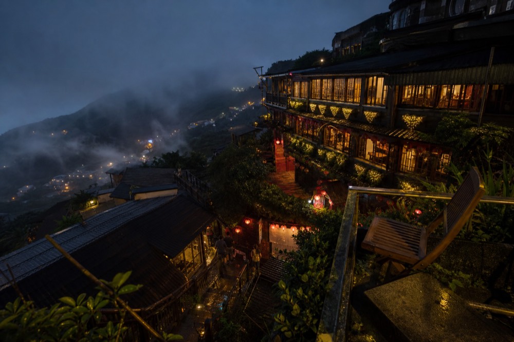 Teahouse windows glowing warm above dark tiled rooftops on a steep hillside at night