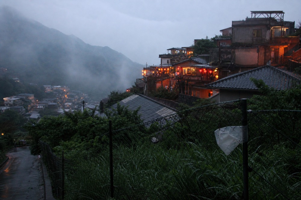 Mist drifting over a hillside town with warm lantern light in the early evening