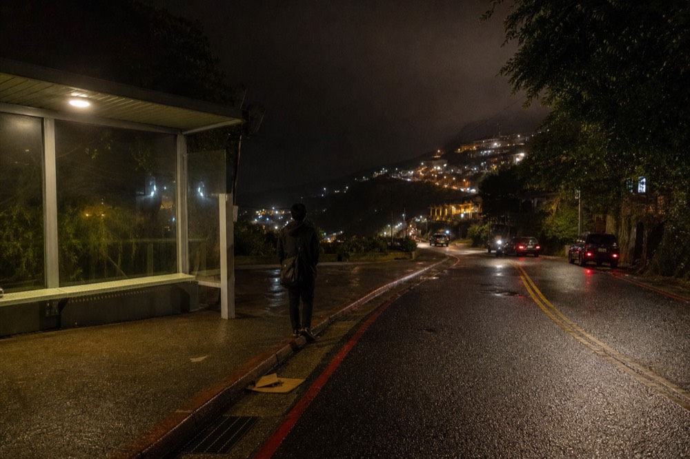 A lit bus stop and quiet road on a hillside at night with town lights below