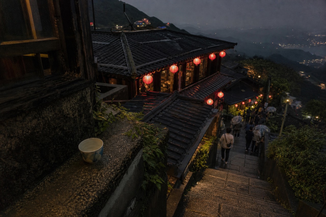 Red lanterns glowing along a narrow stepped street of old teahouses on a hillside at dusk