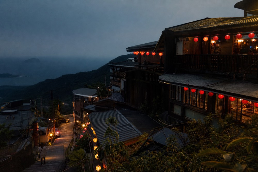 Wooden teahouse balconies and red lanterns on a hillside above a dark sea at dusk