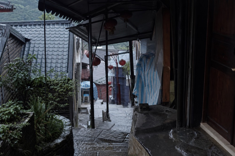 A narrow stone lane in a hillside town with red lanterns hanging overhead and no people