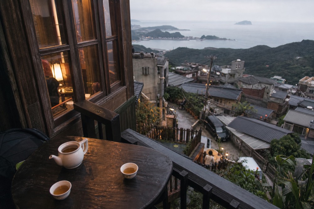 A wooden teahouse balcony with tea sets on the table and a view over rooftops to the sea