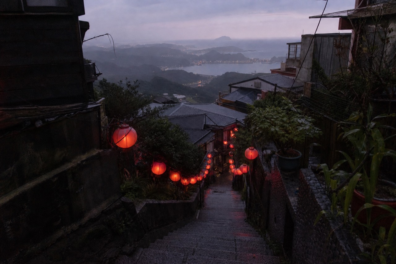 Red lanterns and steep rooftops of a mountain town above the sea at dusk